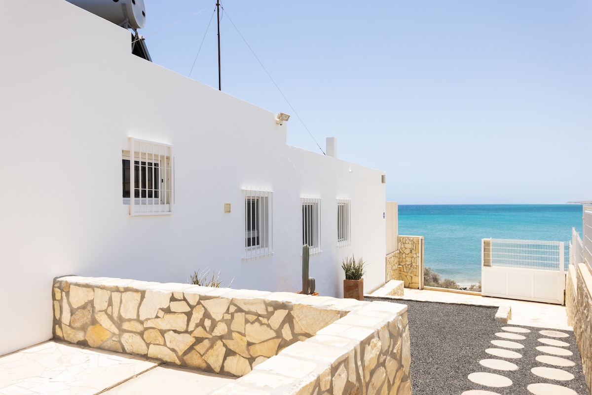 A whitewashed bungalow with multiple windows is set against a serene coastal backdrop. The pathway features smooth stone elements leading to a pebbled courtyard, with the tranquil blue sea visible in the distance, enhancing the sense of peaceful seclusion.