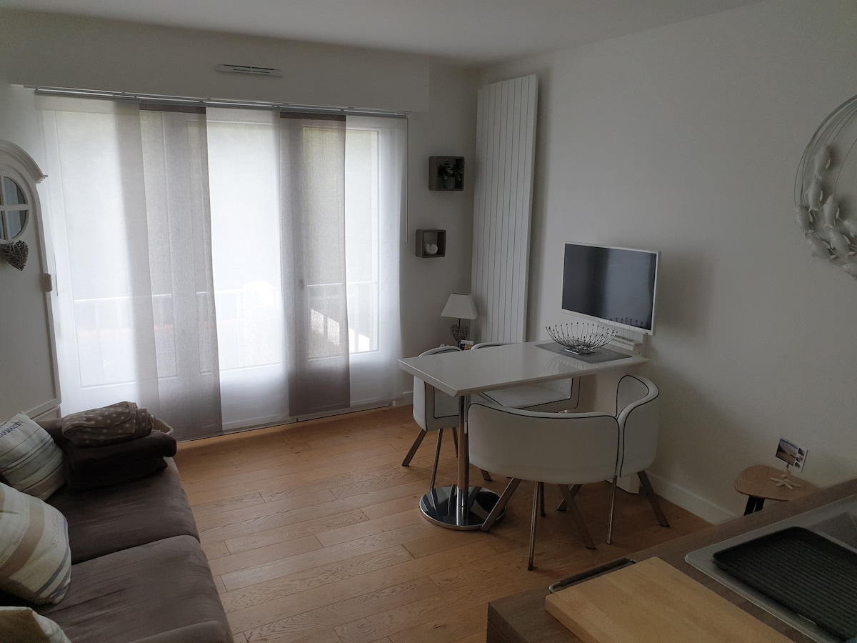 A bright living area features a small work desk with two white chairs positioned beside a window with sheer curtains. A flat-screen television is mounted on the wall, while a comfortable sofa with light-colored cushions invites relaxation. Light wood flooring complements the modern decor.