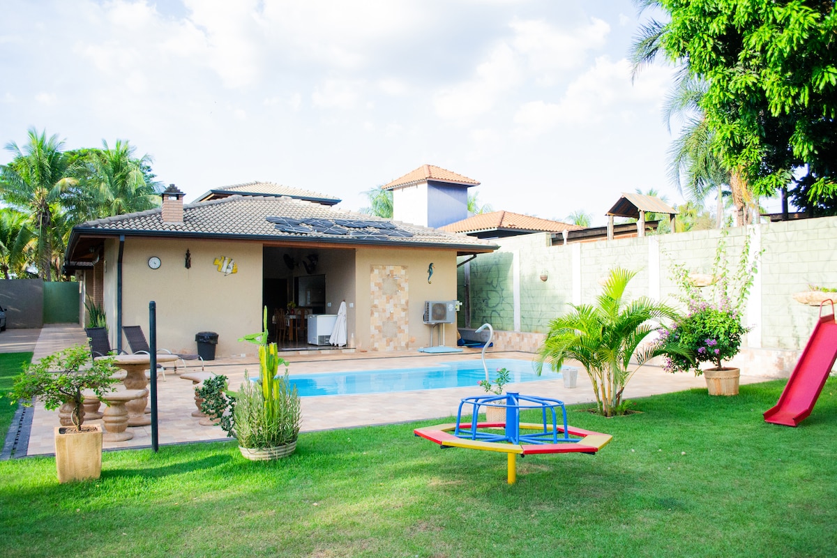 A well-maintained outdoor area features a swimming pool surrounded by a neatly cut lawn. A slide and a colorful playground set are visible, along with lush greenery. The house, with a tiled roof, can be seen in the background, alongside a patio area for relaxation.