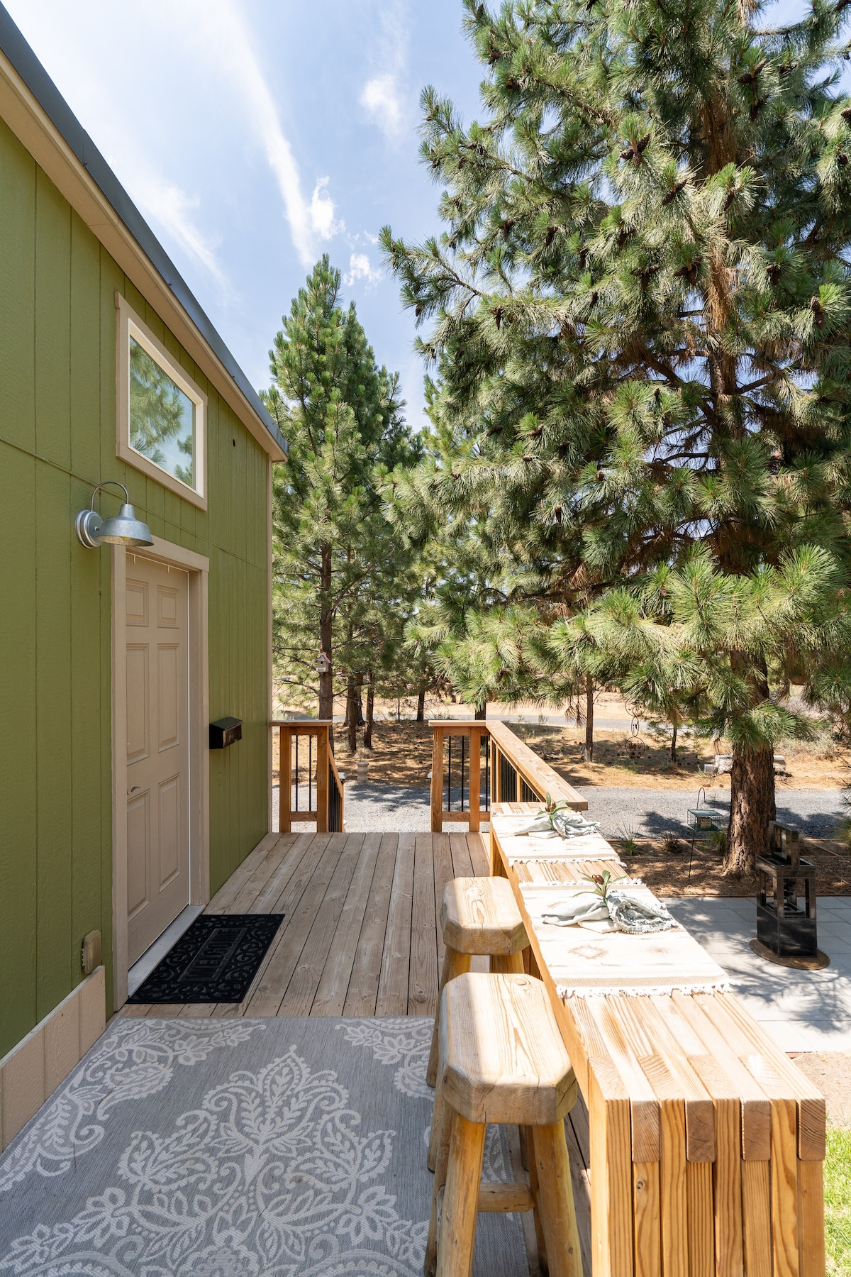 The exterior of the tiny house features a welcoming entrance with a light-colored front door and a wooden deck. A rustic dining area with bar stools is situated beside the house, surrounded by lush pine trees and a manicured lawn.