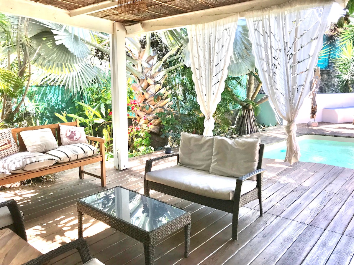 A covered outdoor area features a cozy seating arrangement with a glass table and soft cushions. Tropical foliage surrounds the space, providing a sense of privacy. A glimpse of a tranquil pool can be seen in the background, enhancing the serene environment.