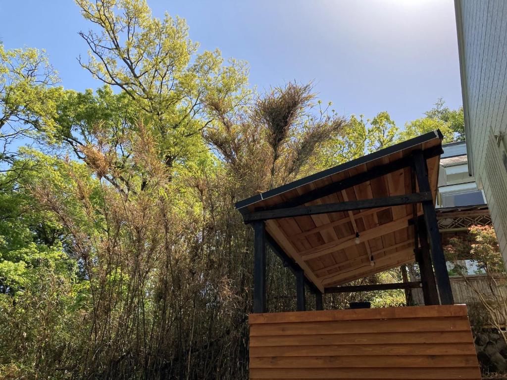 A wooden structure is partially visible among lush greenery, suggesting a blend of nature and shelter. The roof appears to be slanted, offering shade from the sun. Various trees and foliage surround the area, enhancing the natural ambiance.