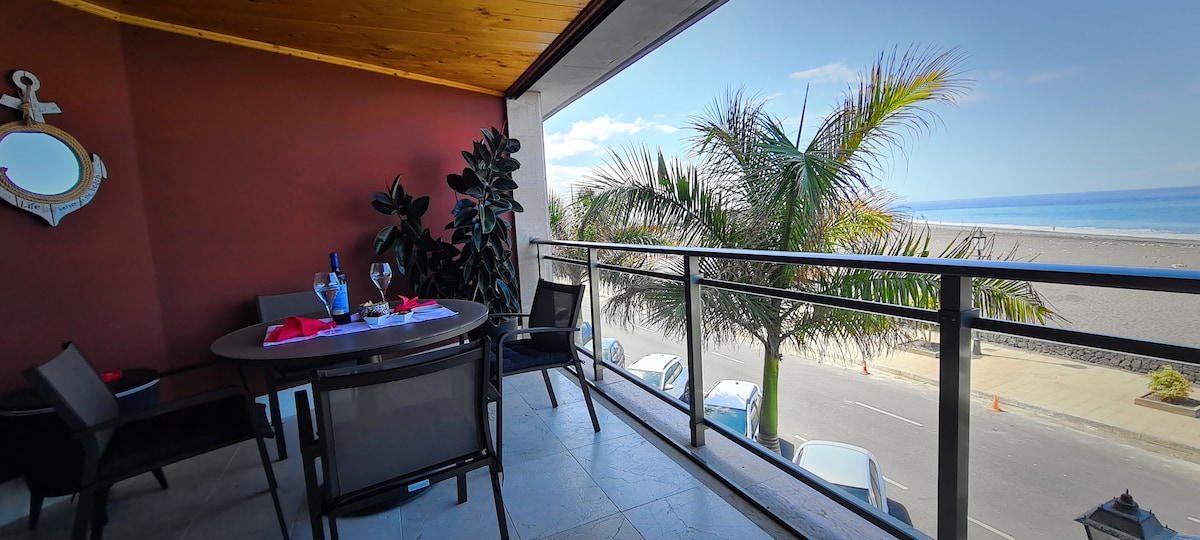 A spacious balcony is visible, featuring a round dining table set with a bottle and glasses. Lush green plants provide a natural touch, while the ocean and beach can be seen in the distance under a clear blue sky.