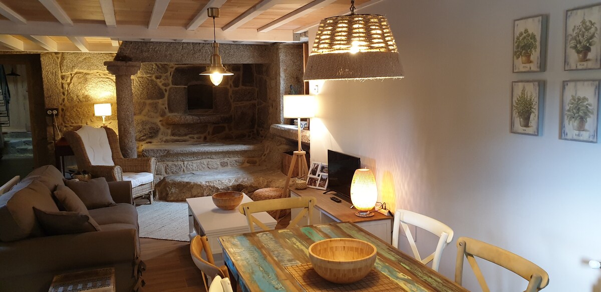 The cozy living area features a rustic stone fireplace, surrounded by soft lighting from various lamps. A wooden dining table with light-colored chairs sits in the foreground, complemented by decorative bowls. The room's design incorporates natural materials, creating a harmonious atmosphere.