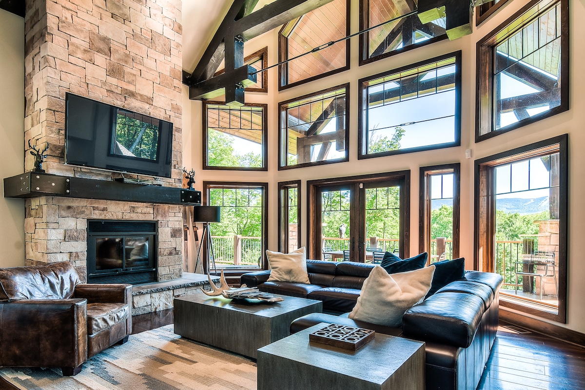 A spacious living room features a stone fireplace as a focal point, complemented by large windows that provide ample natural light and views of the surrounding nature. Comfortable seating is arranged around a modern coffee table, creating a welcoming gathering space.