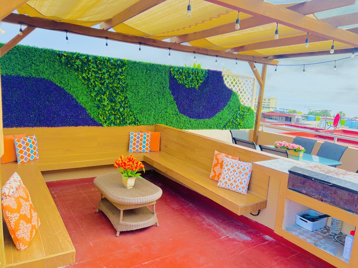 A rooftop terrace is showcased with cozy built-in seating adorned with bright orange and patterned cushions. A central coffee table is presented on terracotta tiles, surrounded by lush green wall accents and string lights overhead, creating an inviting space for relaxation.