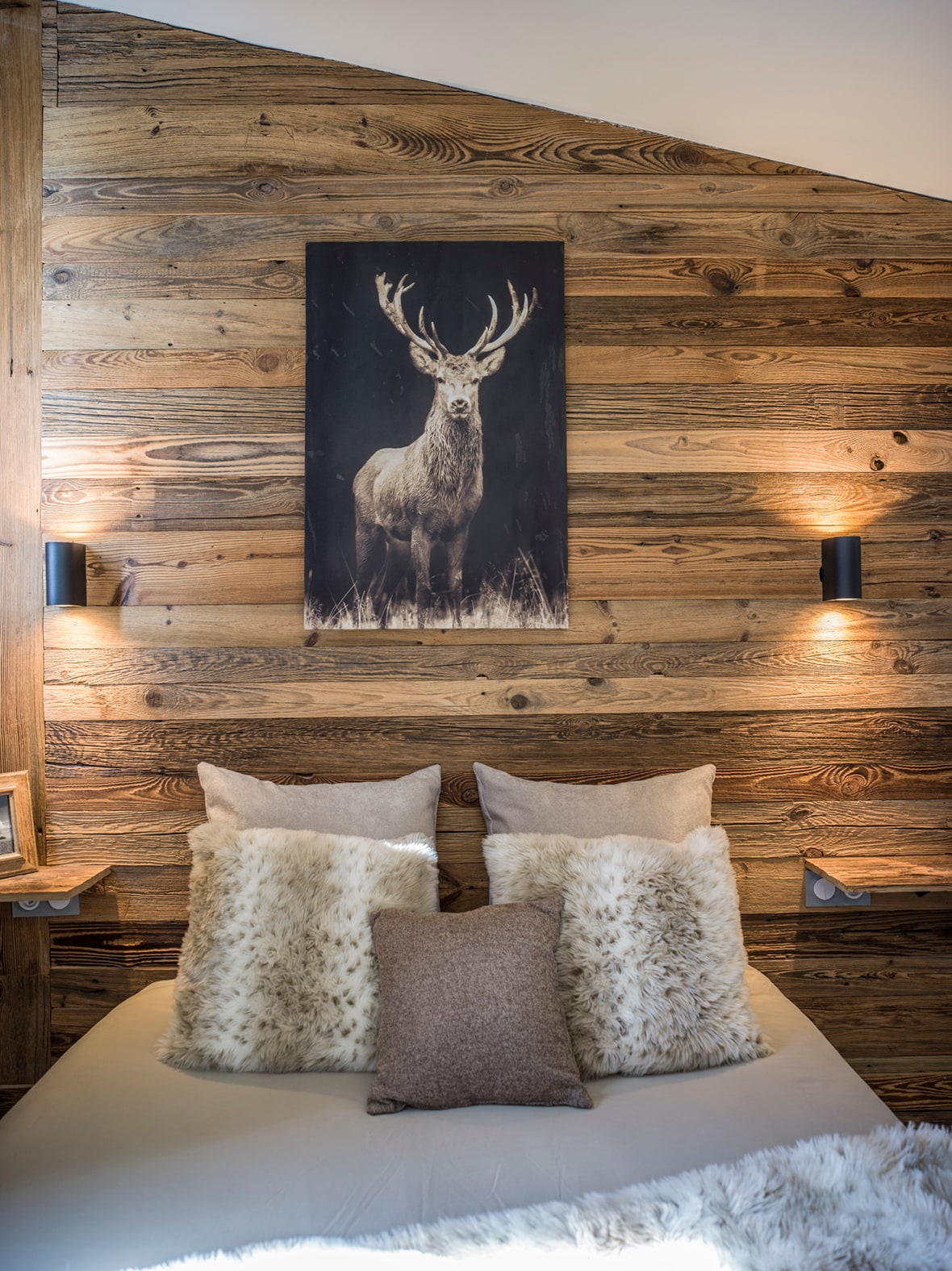A cozy sleeping area is presented with a spacious bed adorned with various decorative pillows. Warm wooden walls create a rustic ambiance, complemented by soft lighting from wall-mounted sconces. A striking artwork of a deer adds a nature-inspired element to the room.