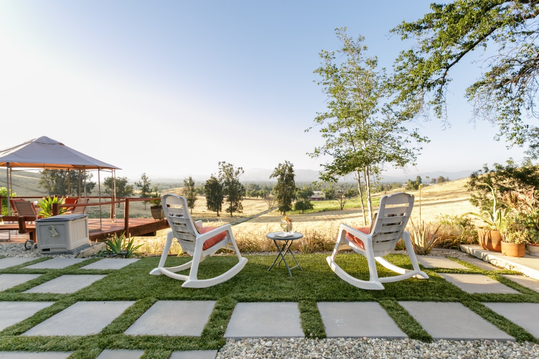 Two white rocking chairs are positioned on a grassy patio, overlooking a landscape of rolling hills. A small round table sits between the chairs. Potted plants frame the area, while a gazebo and deck can be seen in the background.