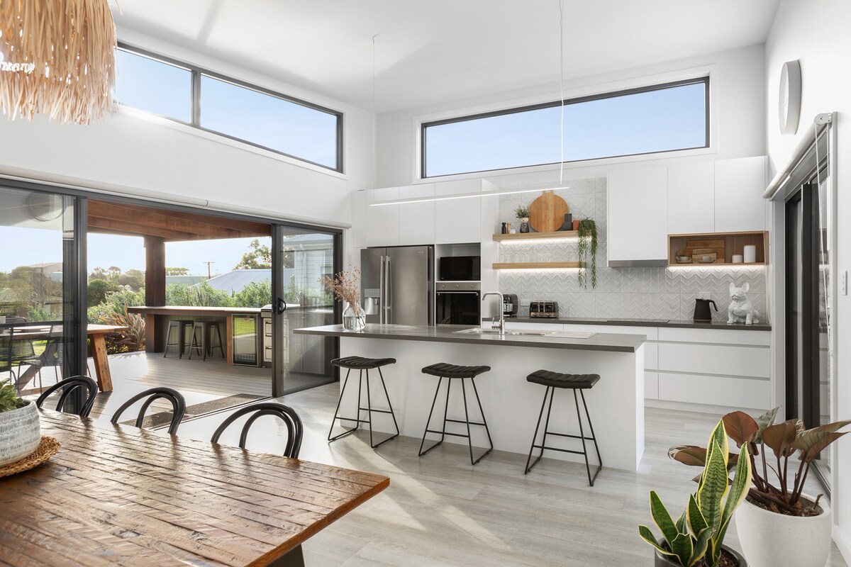 An open-plan kitchen features modern appliances, including a wall oven and microwave. Large windows allow natural light to fill the space, while views of the outdoors and access to a patio are visible. Bar stools are placed at the kitchen counter, complemented by a rustic dining table.