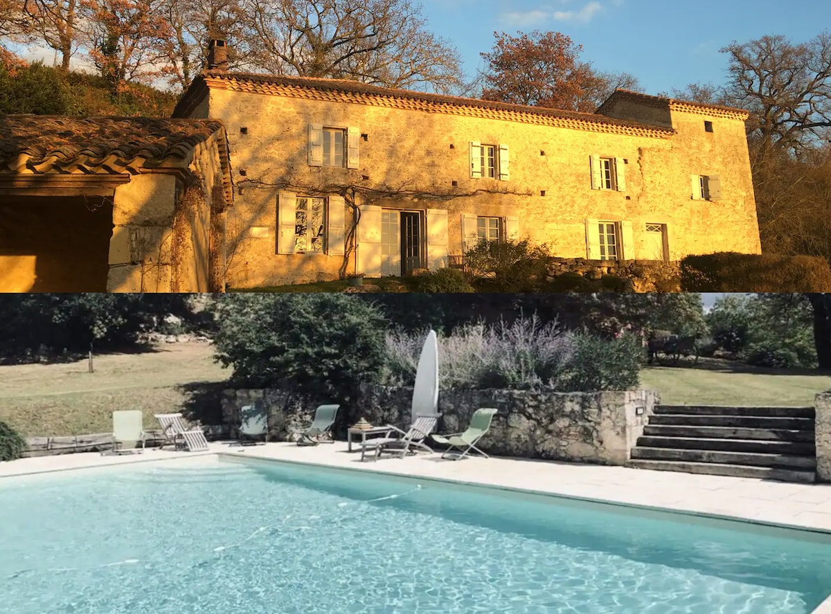 The image showcases a rustic stone building with multiple windows, surrounded by trees. The foreground features a swimming pool, with sun loungers arranged nearby. Gentle steps lead from the pool area to the lush garden, emphasizing a serene outdoor environment.