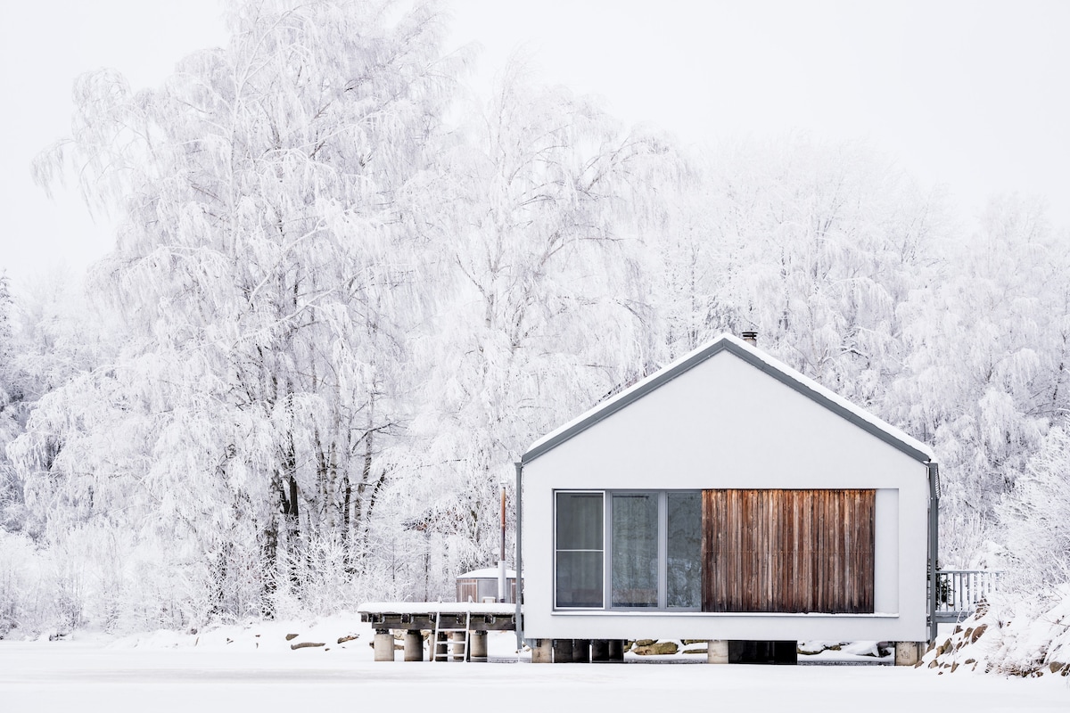 A contemporary house, surrounded by a winter landscape, features a sleek exterior with a combination of white and wooden elements. Large windows reflect the snowy surroundings, while a deck extends out toward the frozen water, inviting views of the serene nature.