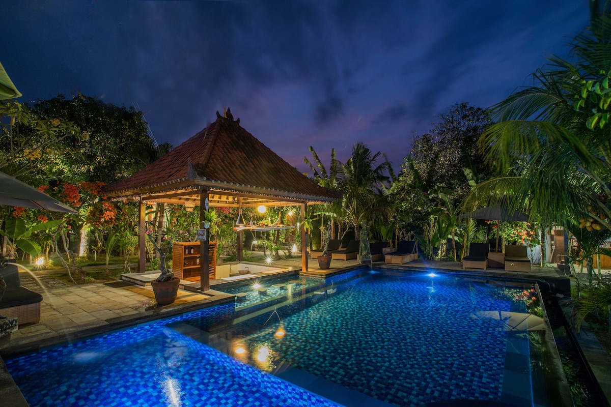 A private pool area is surrounded by lush greenery and garden features. A traditional pavilion offers a shaded seating space beside the pool, which is illuminated by soft lighting. Comfortable lounge chairs are positioned for relaxation under the evening sky.