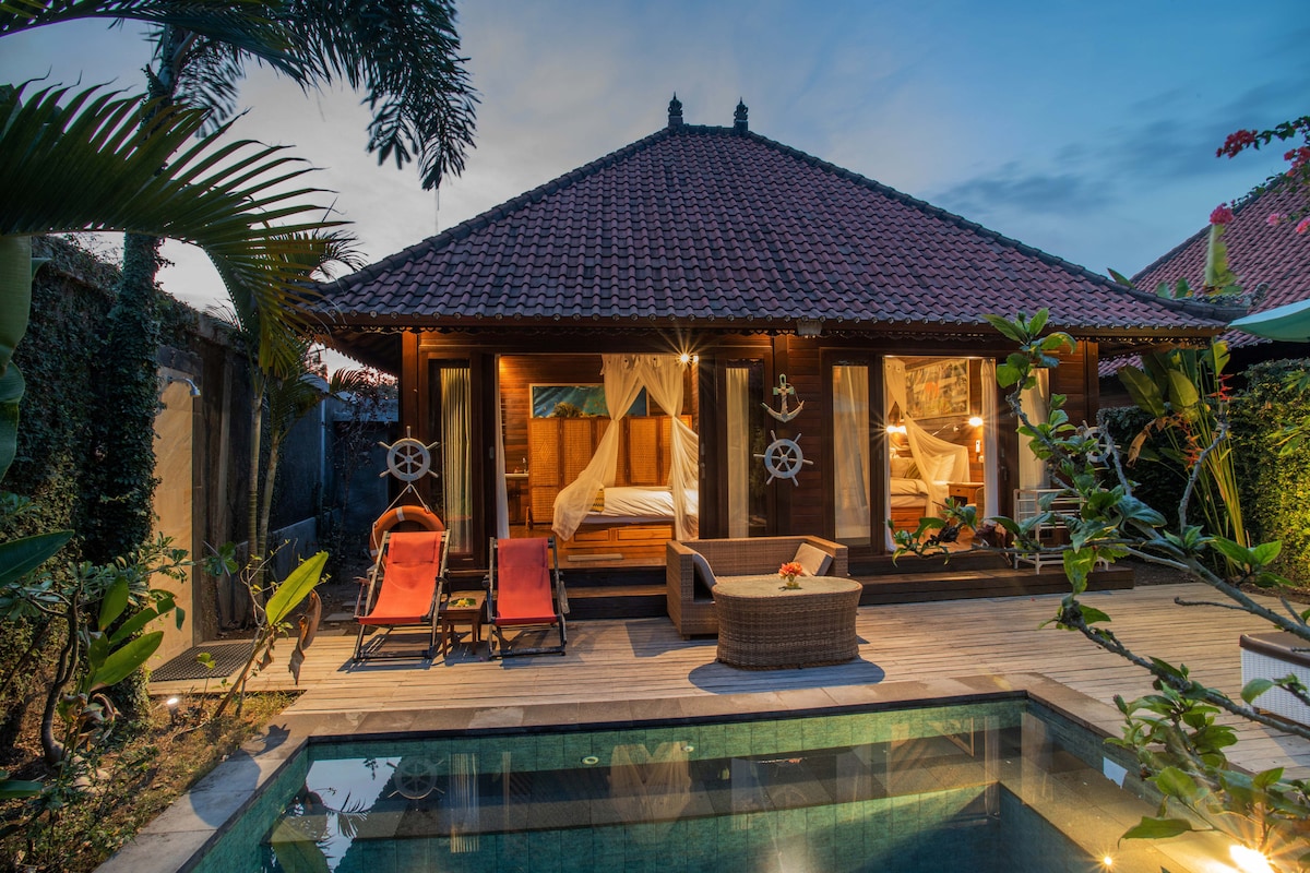 A private villa is shown with a traditional roof, surrounded by lush greenery. A small pool is visible in the foreground, while two lounge chairs sit on a wooden deck. The main entrance reveals a cozy sleeping area with natural light spilling in.