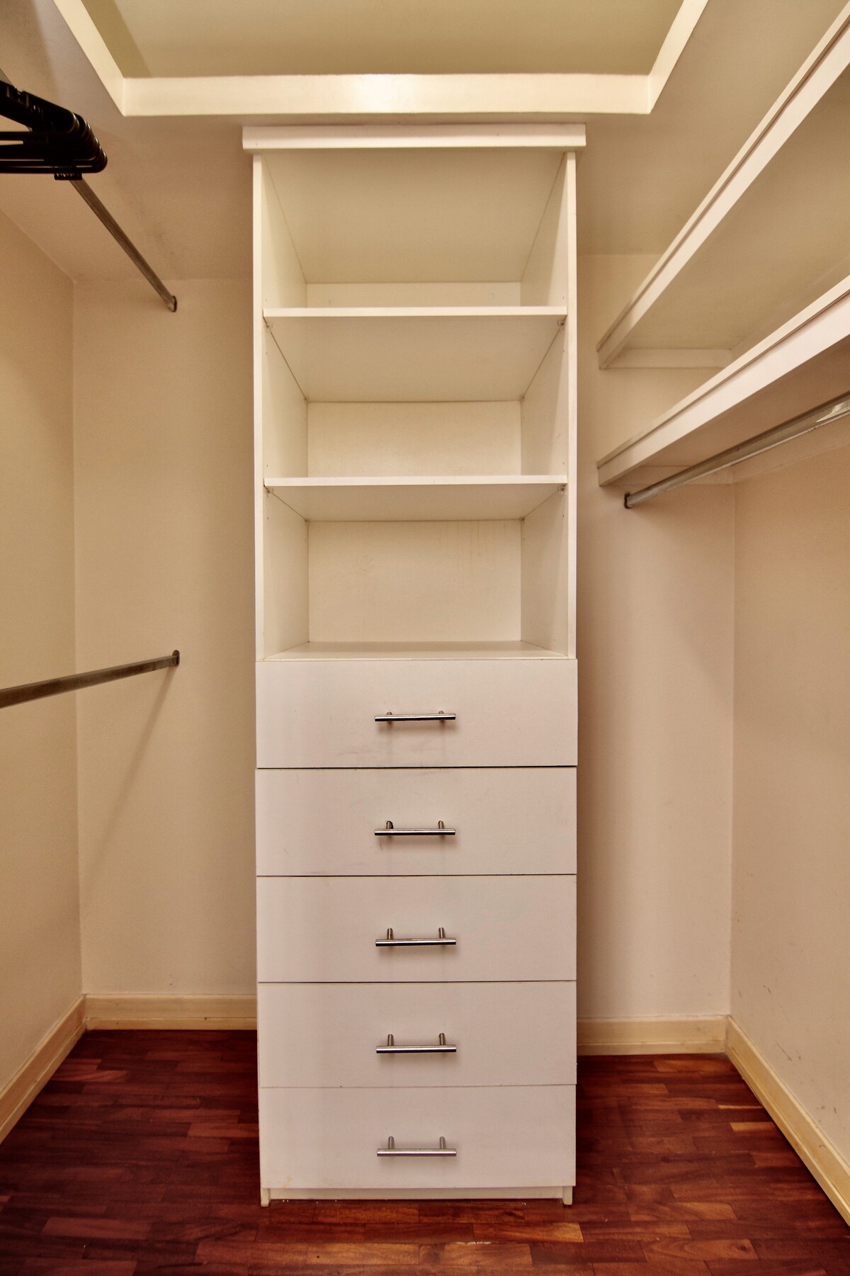 A well-organized closet features a tall built-in shelving unit with five drawers and open shelves above. The wooden floor contrasts with the white cabinetry, while a metal rod for hanging clothes complements the minimalist design.