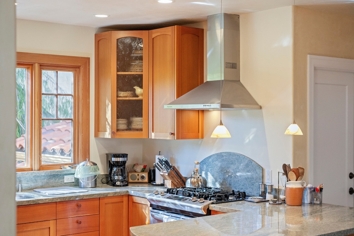 A modern kitchen features warm wood cabinetry with glass doors. Stainless steel appliances, including a stove and coffee maker, are arranged for convenience. A grey stone countertop provides ample workspace, with two pendant lights illuminating the area. Natural light enters through multiple windows.