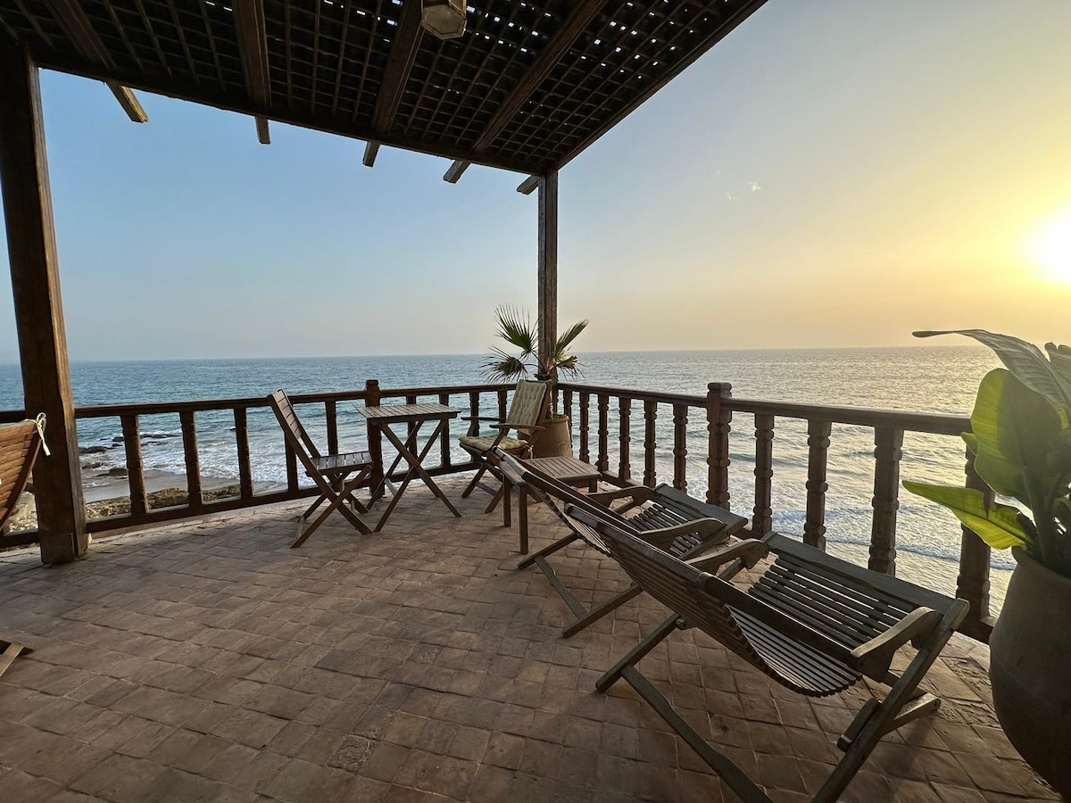 A spacious outdoor terrace extends over the seafront, featuring several lounge chairs and a small table. A potted plant adds a natural element, while the sun sets over the tranquil Atlantic Ocean, casting a soft glow on the surrounding area.