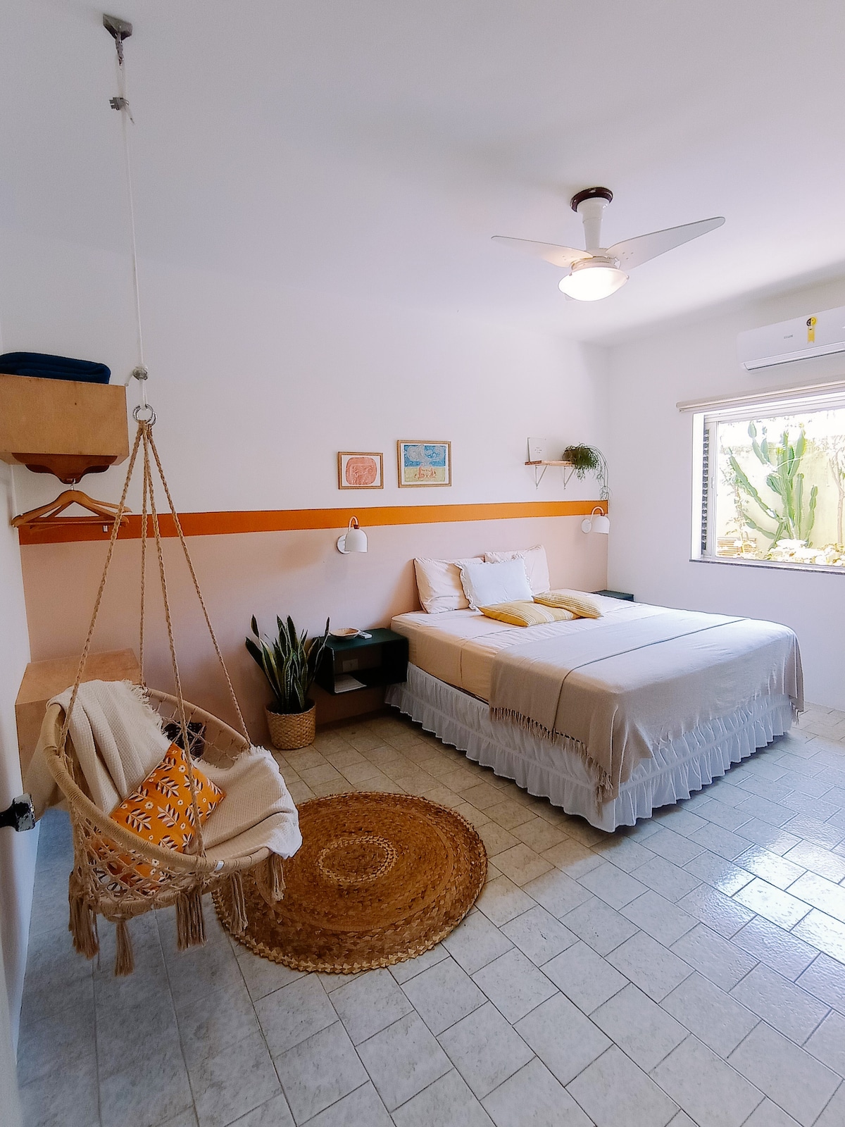 A bright room is equipped with a king-size bed dressed in soft linens. A cozy hanging chair and a small table are positioned beside the bed. Natural light filters in through a window adorned with simple curtains, while a decorative rug adds texture to the tiled floor.