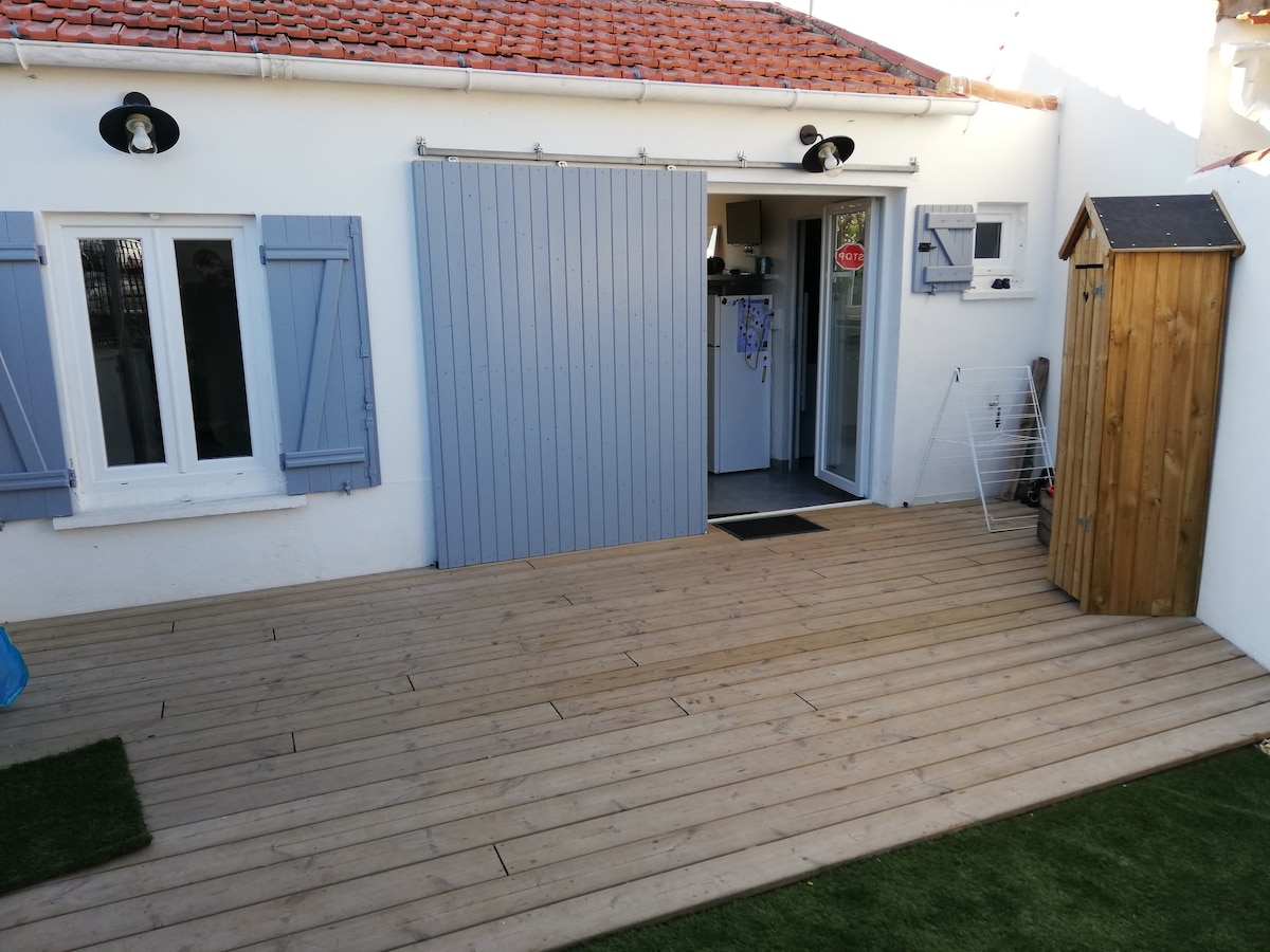 A wooden deck area is shown, featuring a sliding door that leads to an interior space. The walls are painted white, complemented by gray wooden shutters on two windows. A small wooden storage unit stands next to an outdoor drying rack, adding functionality to the area.