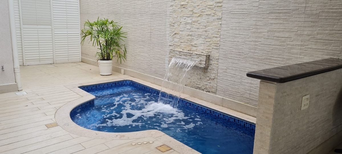 A tranquil spa area features a small, circular pool with a soothing waterfall. The surrounding space is complemented by modern tilework and a potted plant, creating a serene atmosphere. Light reflects off the water, enhancing the inviting ambiance.
