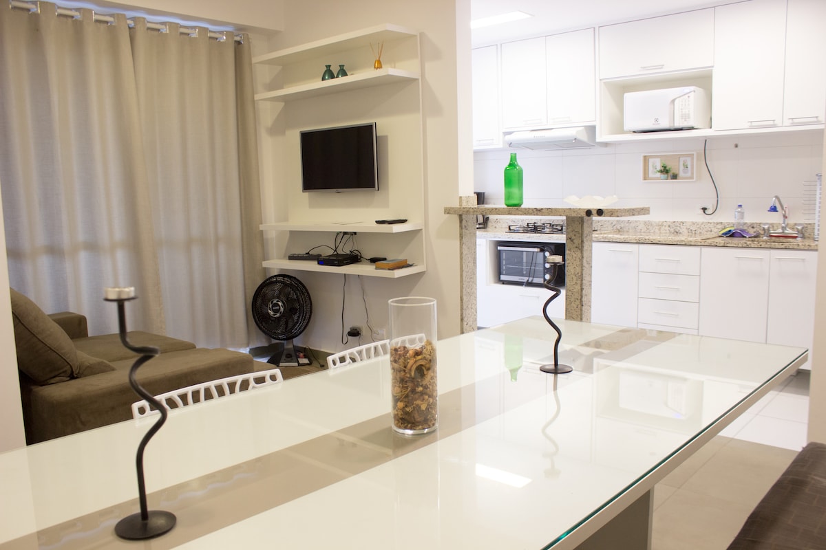 A modern living space is showcased with a well-lit kitchen and dining area. A sleek glass table is set with a decorative vase, while a comfortable sofa sits nearby. The kitchen features contemporary appliances and a clean countertop with utensils visible.