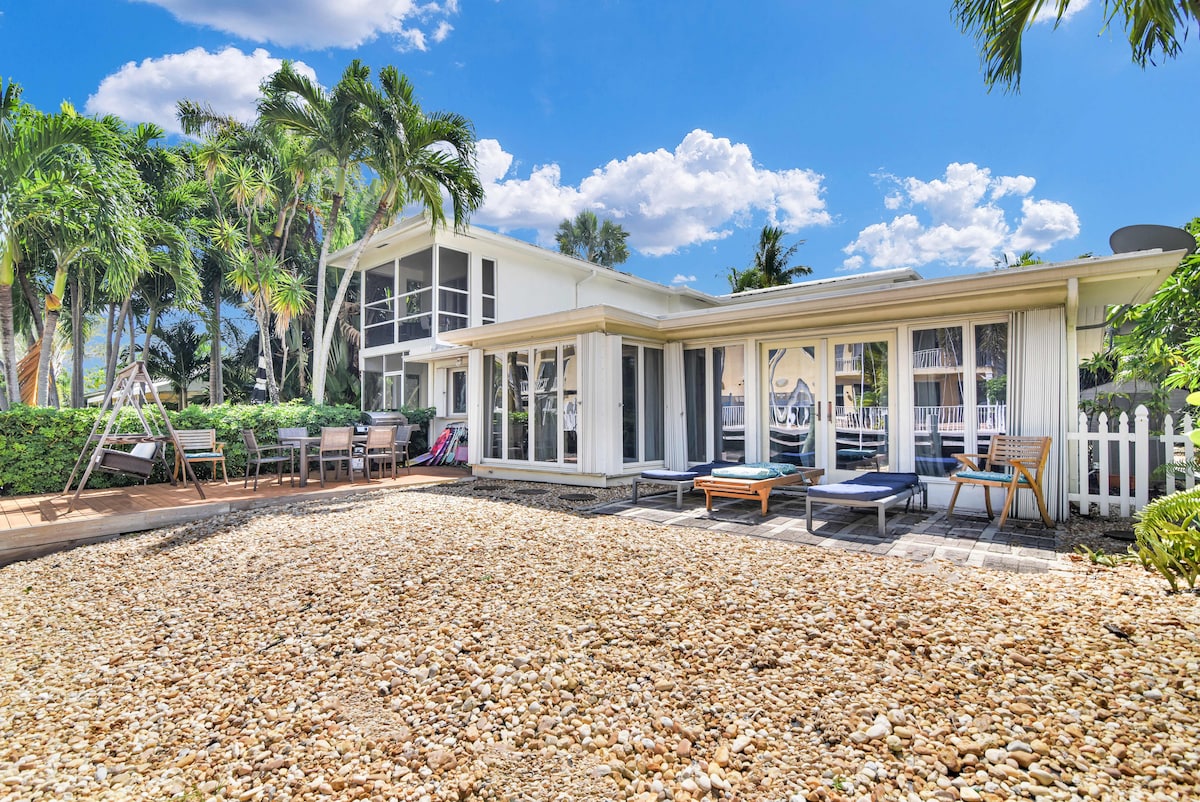 The exterior of the condo showcases a spacious patio area lined with smooth pebbles and surrounded by lush tropical landscaping. Sun loungers are positioned for relaxation, while a dining set is visible in the background. The structure features large glass doors, allowing light to enter.