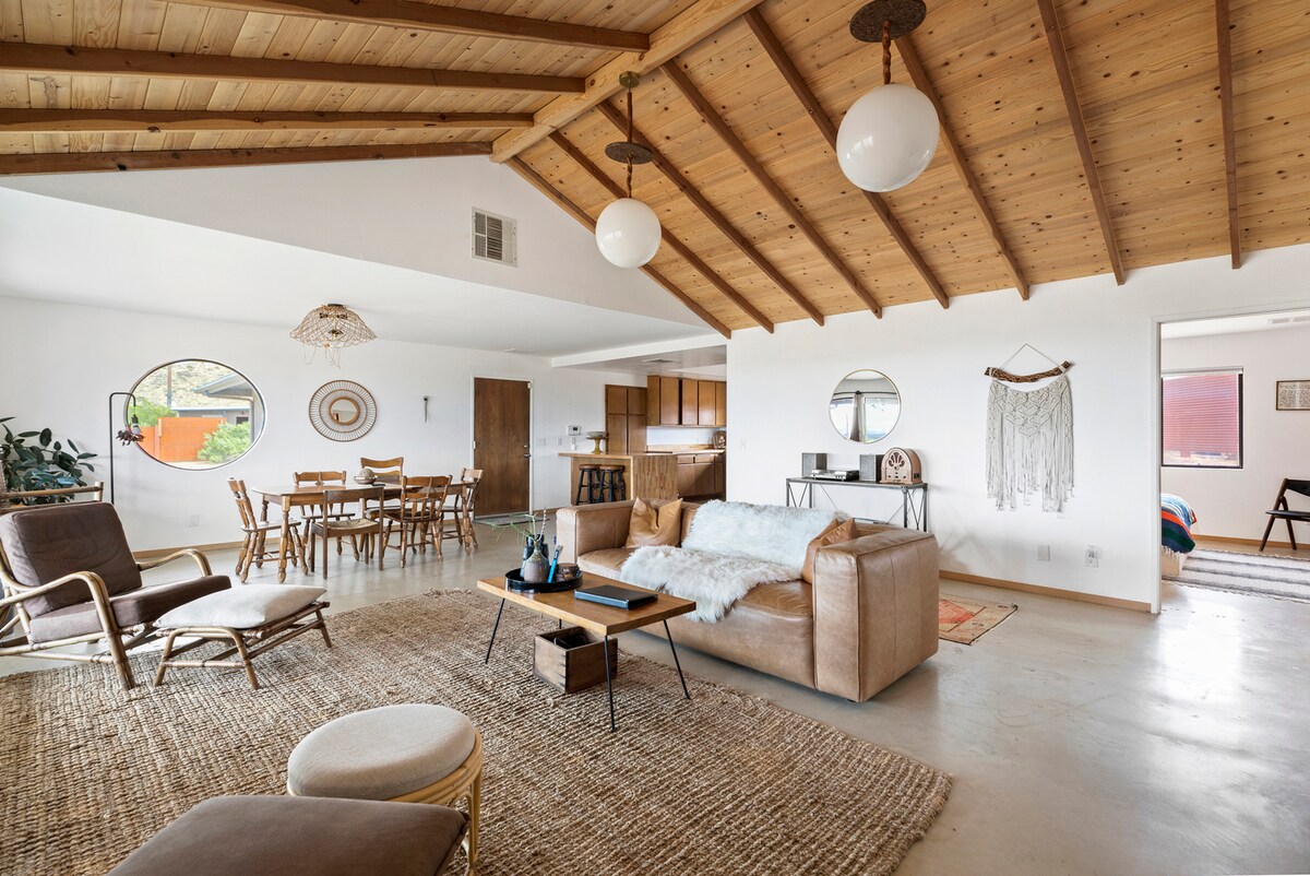 An open main room showcases a vaulted wooden ceiling and a leather sofa. Circular windows provide natural light, while the dining area features a table for six. Earthy textures and neutral tones are complemented by a woven rug, enhancing the space's cozy atmosphere.