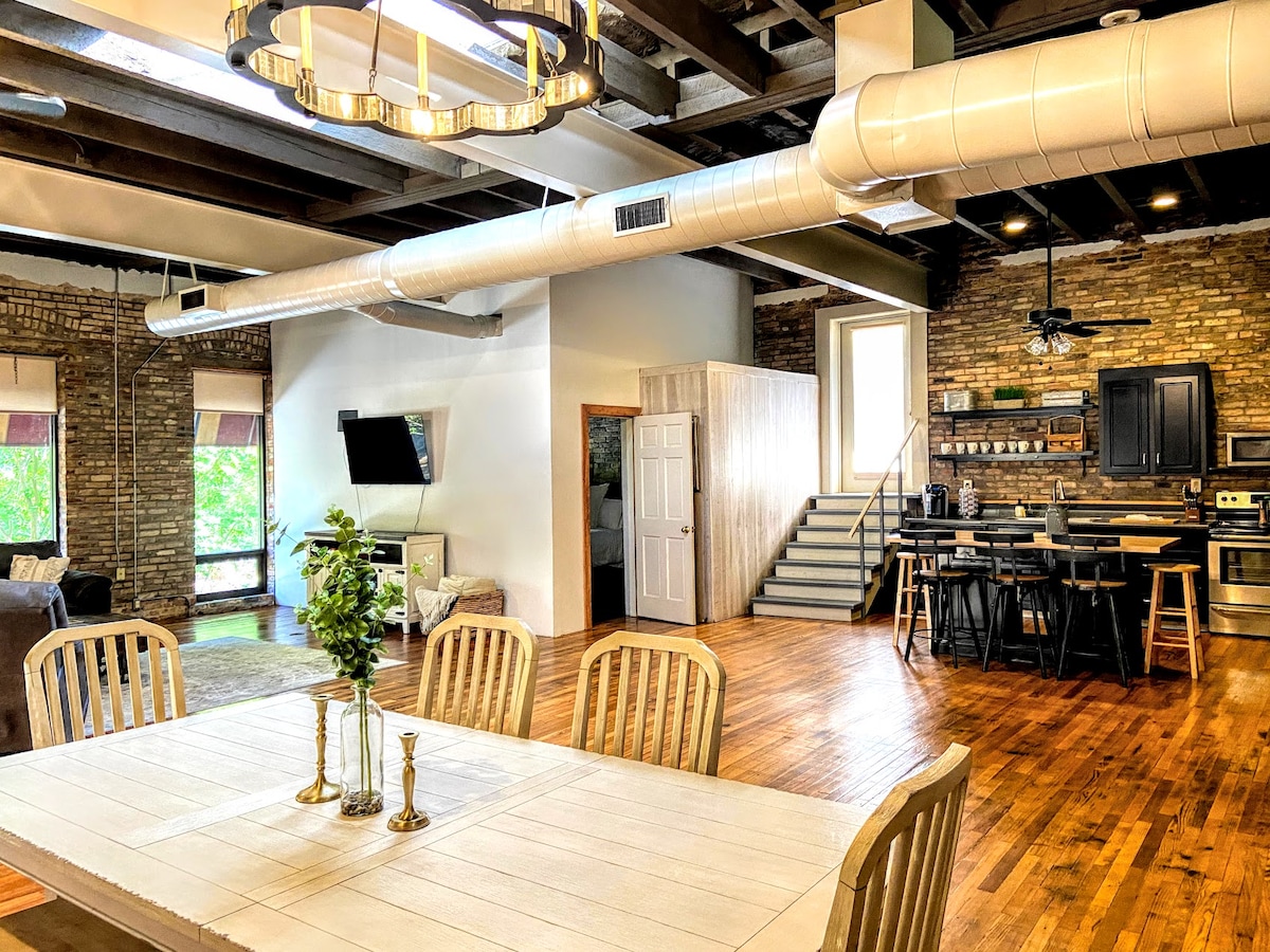 An open living space showcases high ceilings with exposed beams. A wooden dining table is surrounded by light-colored chairs, while a modern kitchen features dark cabinetry and stainless steel appliances. Natural light floods in through large windows, highlighting the exposed brick walls.