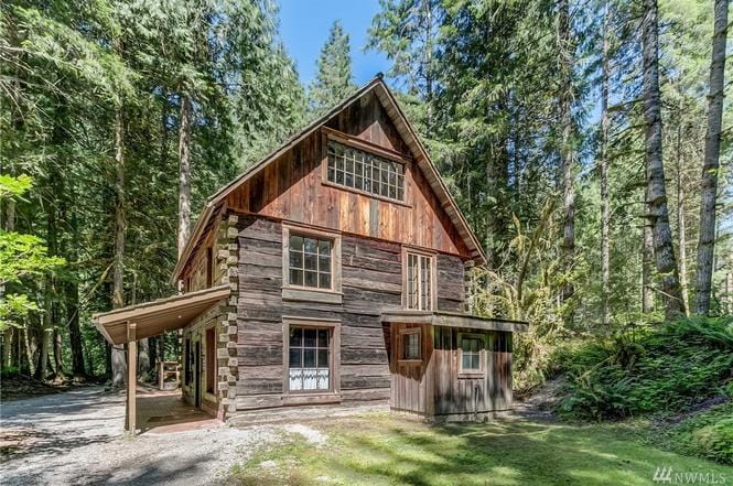A rustic wooden cabin is nestled among dense coniferous trees. Its charming architecture features a sloped roof, multiple windows allowing natural light, and a combination of weathered brown and gray timber accents. A gravel path leads up to the structure.