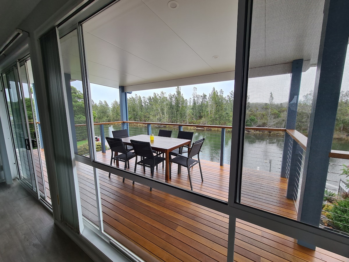 A spacious outdoor balcony features a wooden table surrounded by chairs, offering views of the tranquil water and lush greenery. Large glass doors enhance natural light and create a seamless connection between indoor and outdoor spaces.