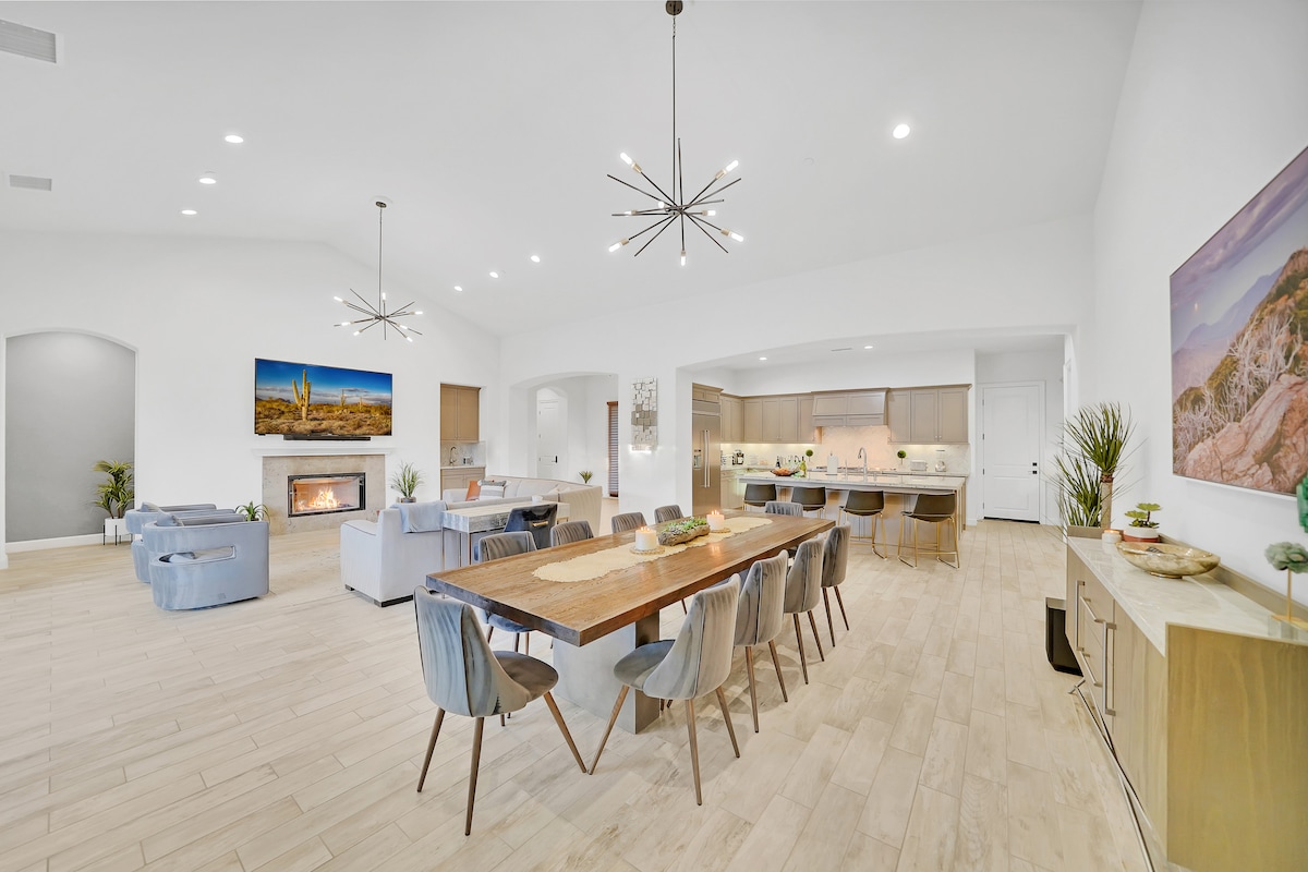 A spacious great room is seen with high vaulted ceilings and modern lighting fixtures. The dining table accommodates multiple guests, surrounded by stylish chairs. A cozy sitting area is positioned near a fireplace, alongside sleek cabinetry and contemporary decor.