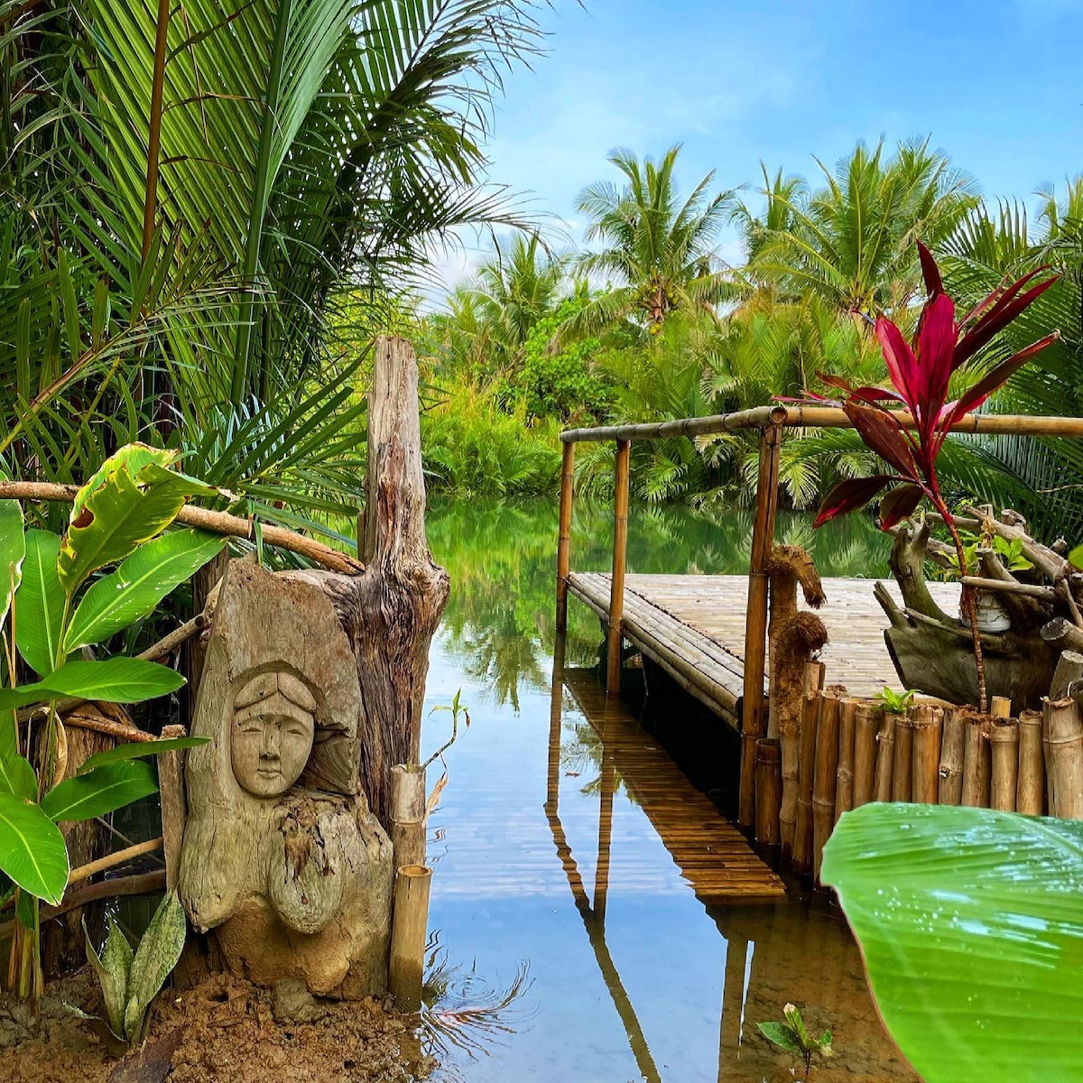 A serene wooden deck extends over calm waters, surrounded by lush greenery. Tropical plants and vibrant red foliage frame the scene, creating a tranquil atmosphere. Reflections of palm trees can be seen on the water's surface, enhancing the natural beauty of this oasis.