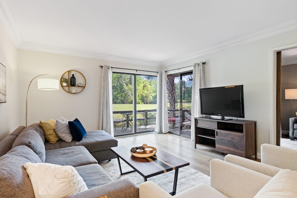 A spacious living area features a large sectional sofa complemented by soft cushions, along with two light armchairs. Floor-to-ceiling sliding glass doors offer views of the lush exterior. A television is placed on a modern wooden stand, enhancing the contemporary design.