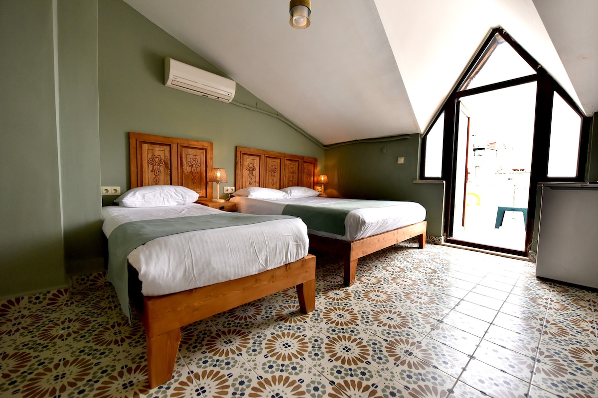 A spacious room featuring two beds with wooden frames, positioned on a tiled floor with a decorative pattern. Large windows extend to the ceiling, providing natural light and a view of the surroundings. An air conditioning unit is mounted on the wall for comfort.