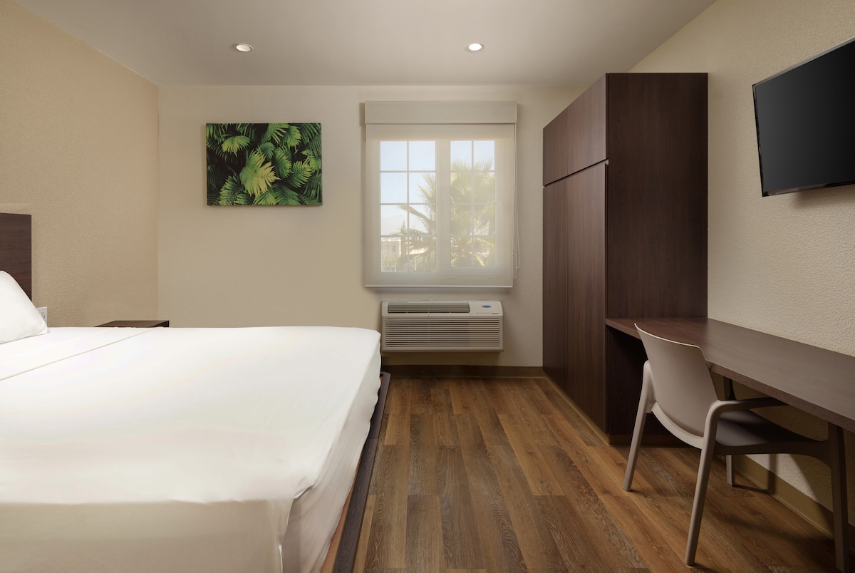 A bright guest room features a spacious bed with crisp white linens and a window offering natural light. A mounted television is visible on the wall opposite. A wooden desk with a chair and a storage cabinet complement the simple, modern design.
