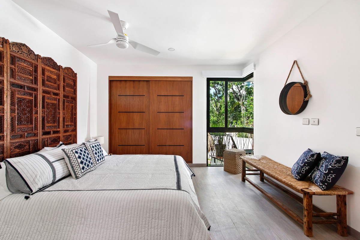 A spacious bedroom features a large bed with a soft quilt and decorative pillows. A wooden wardrobe with a textured design is visible, while a cozy bench made of natural materials is positioned nearby. Large windows provide views of the surrounding lush greenery.