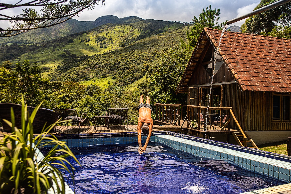 A swimmer is captured diving into a serene blue pool, surrounded by lush greenery and mountainous landscapes. A wooden deck with seating is visible in the background, complementing the natural setting.