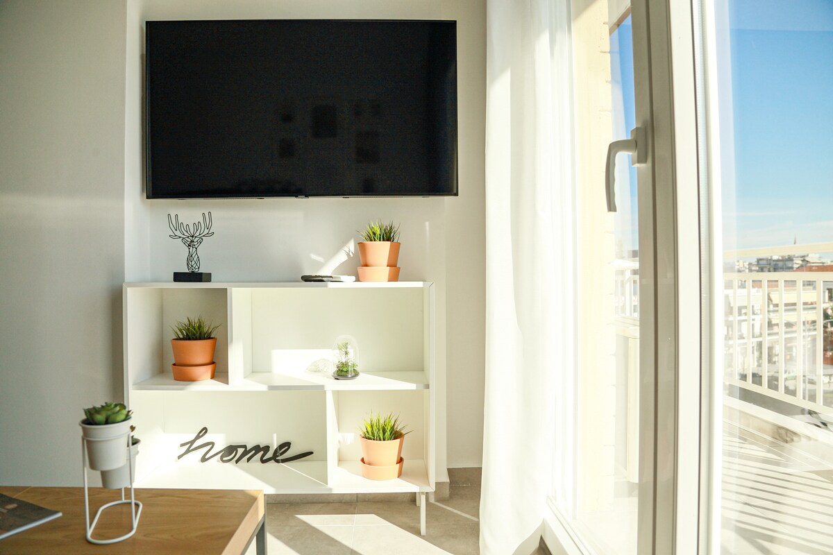A modern living area features a large wall-mounted television above a stylish white media console. Small potted plants decorate the shelves, while natural light floods the space through a nearby window, highlighting the clean lines and minimalist decor.