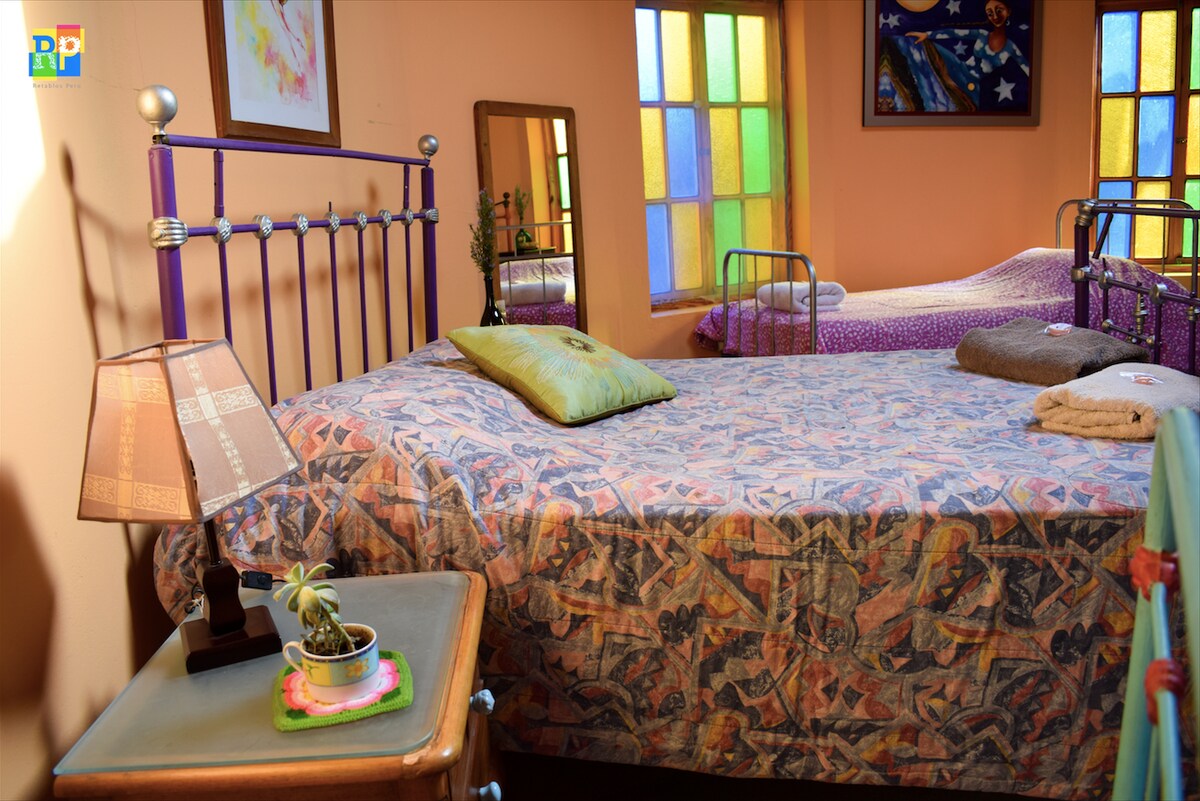 A spacious sleeping area features a patterned bedding set atop a metal bed frame. Two additional beds are positioned in the background, each with individual covers. Colorful stained glass windows allow soft light to illuminate the room, complemented by a bedside lamp and a small potted plant.
