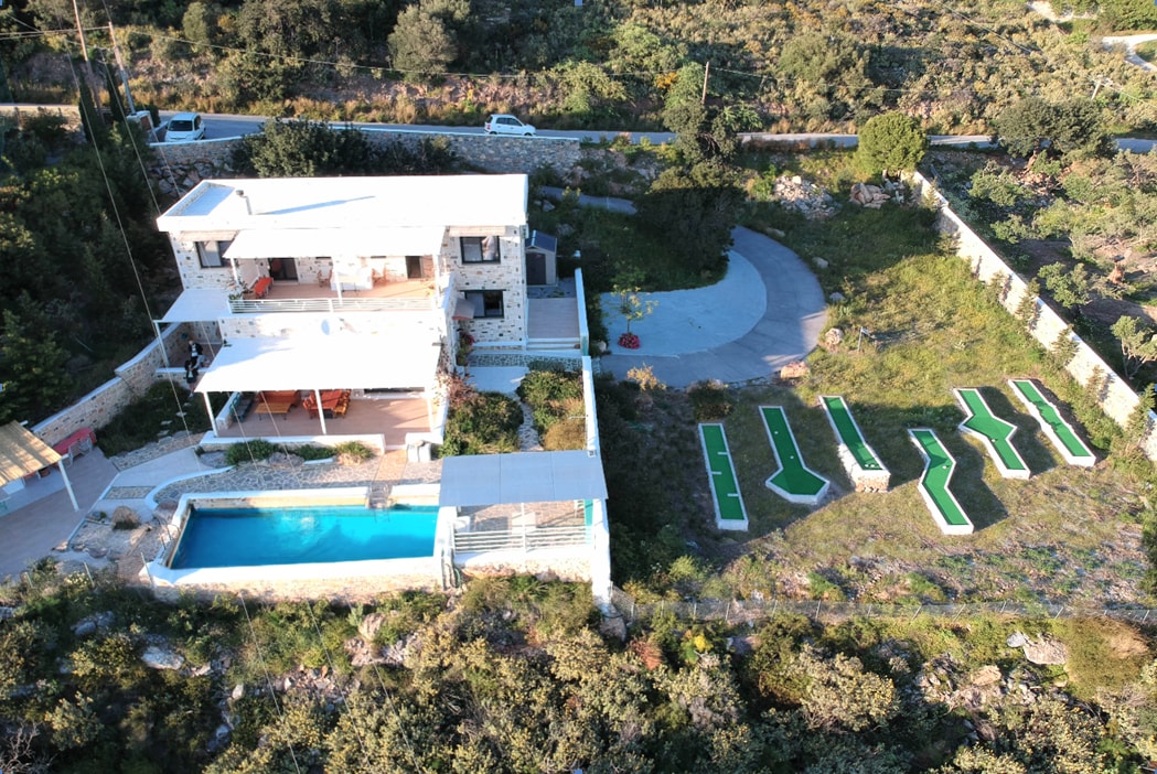 An aerial view of Villa Anasa displays a modern structure surrounded by lush greenery. A spacious salt-water pool is positioned at the front, while a circular driveway is visible alongside. The serene landscape is dotted with wild herbs and features a well-maintained outdoor space.