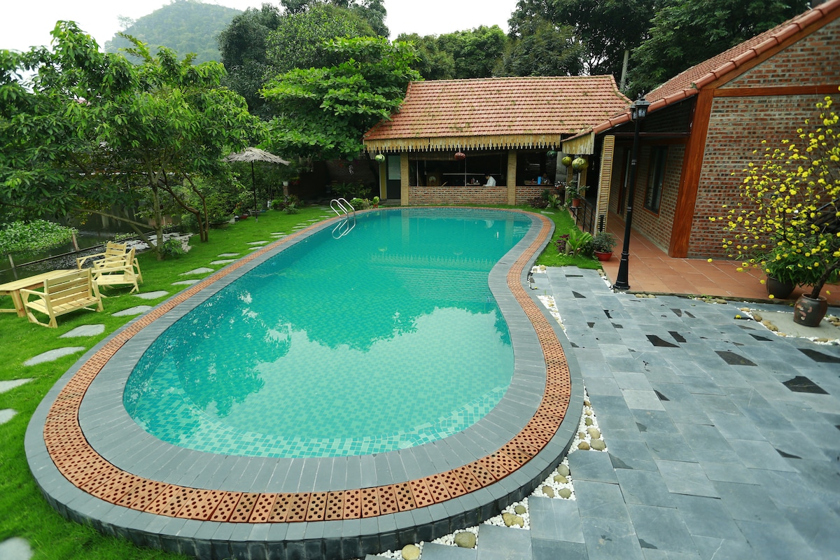 A serene garden area features a curved swimming pool surrounded by lush greenery. The pool is bordered by a patterned stone walkway, and a shaded seating area can be seen nearby. Trees provide a natural backdrop, enhancing the peaceful outdoor setting.