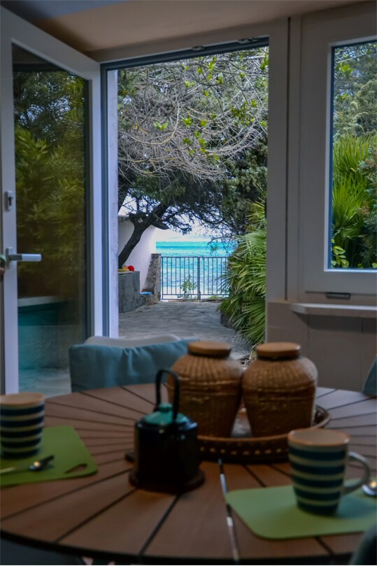A dining area is seen through large windows, framed by greenery and overlooking the sea. A round wooden table is set with two striped cups and placemats. Two woven baskets sit in the center, creating a welcoming atmosphere.