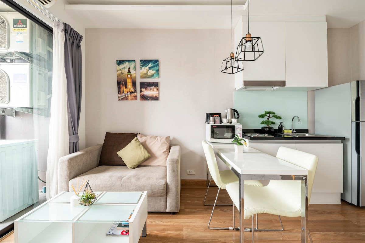 A modern living space features a comfortable sofa adorned with a decorative pillow, a dining table with two chairs, and a well-equipped kitchen area. Artwork in simple frames adds color to the wall, while natural light fills the room through the balcony doors.