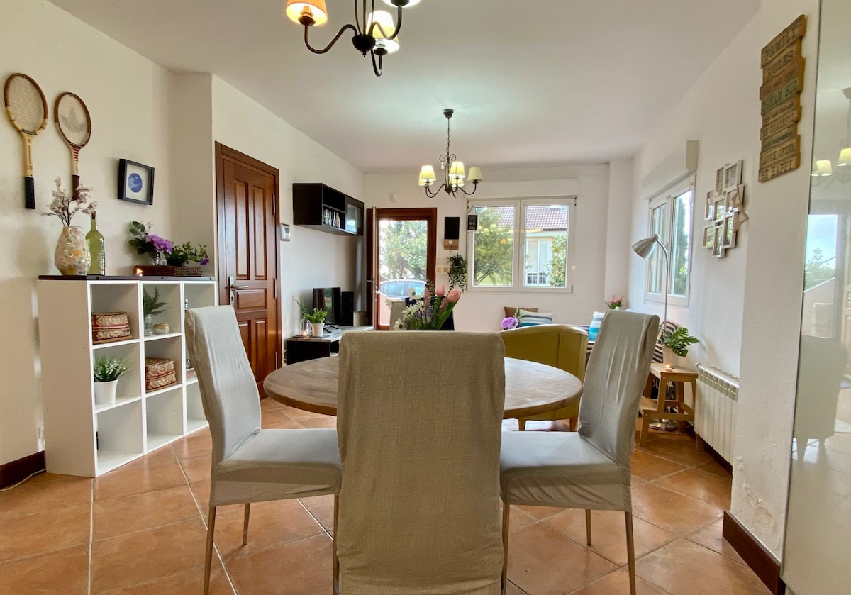 A bright living area features a round dining table surrounded by light-colored chairs. Natural light fills the space through multiple windows, highlighting the modern decor that includes a television and decorative shelving units. A welcoming atmosphere is enhanced by greenery and tasteful furnishings.