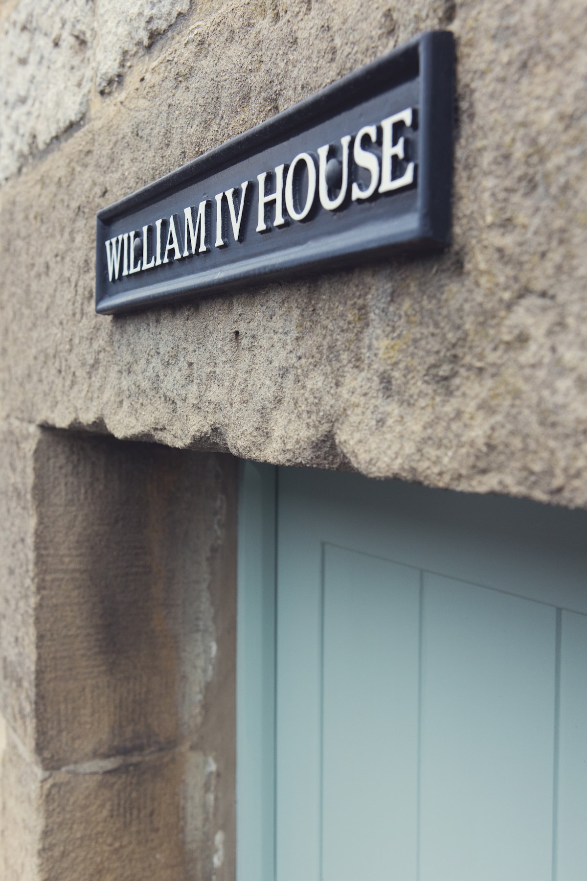A rustic stone facade features a sign reading 'William IV House' positioned above a pale blue door. The texture of the stone is visible, showcasing its historic charm.