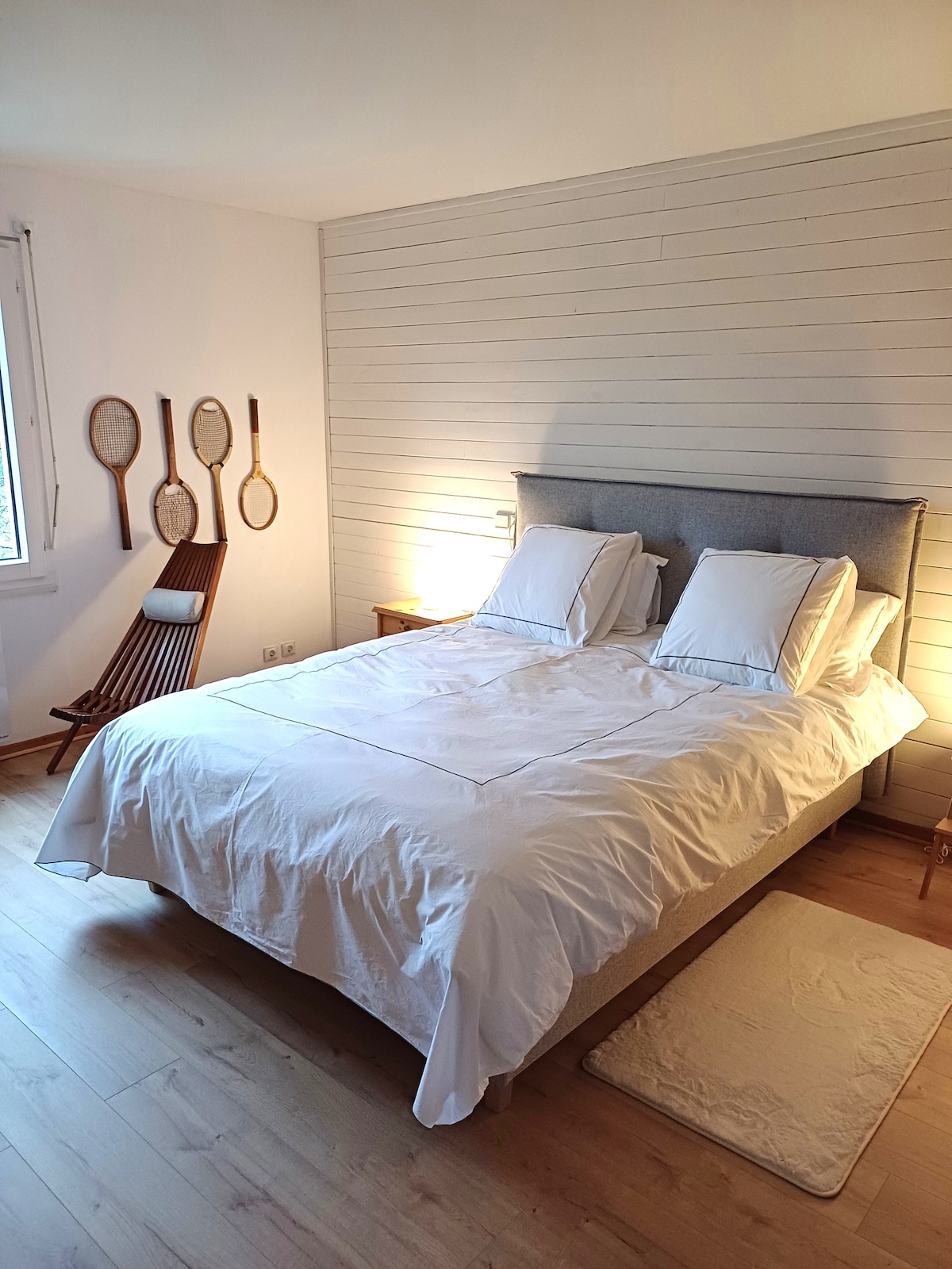 A bright sleeping area features a spacious bed with white linens and two pillows. A wooden chair and four tennis rackets are displayed on the wall, and soft lighting emanates from a bedside lamp, creating a calm environment.