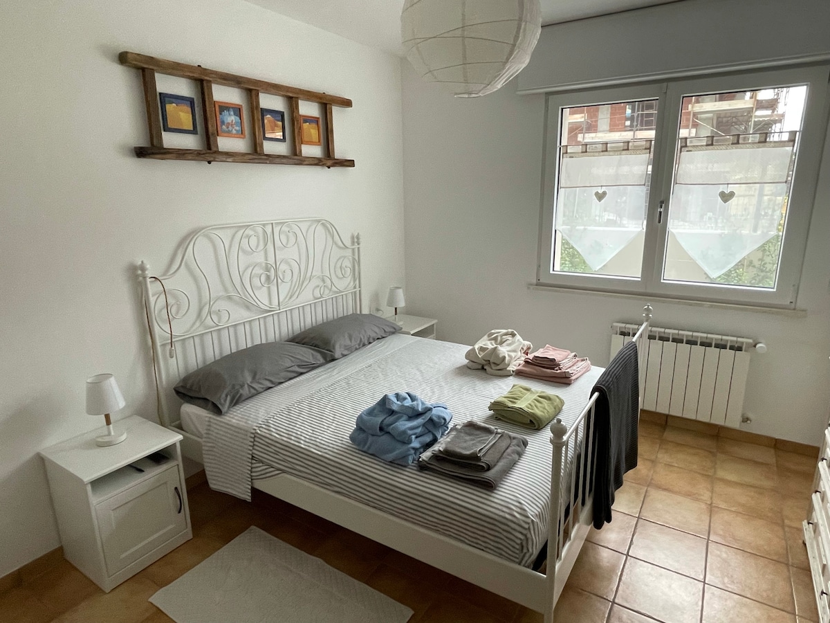 The sleeping area features a light-colored metal bed with neatly arranged linens and towels in various colors on top. Two bedside tables with lamps flank the bed. A large window allows natural light to enter, while framed artwork hangs above the headboard.