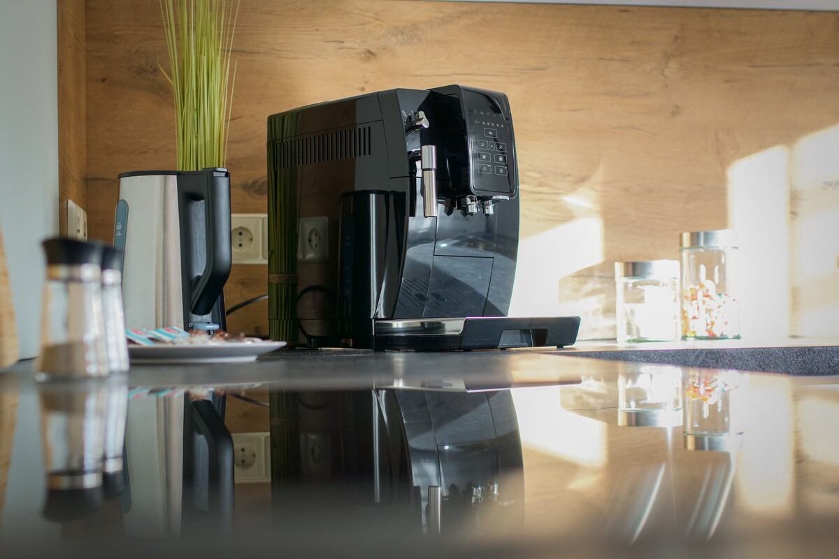 A coffee machine is prominently displayed on a sleek countertop, reflecting natural light. A kettle and a jar filled with items are visible in the background, alongside a salt and pepper shaker set, enhancing the functional kitchen space.