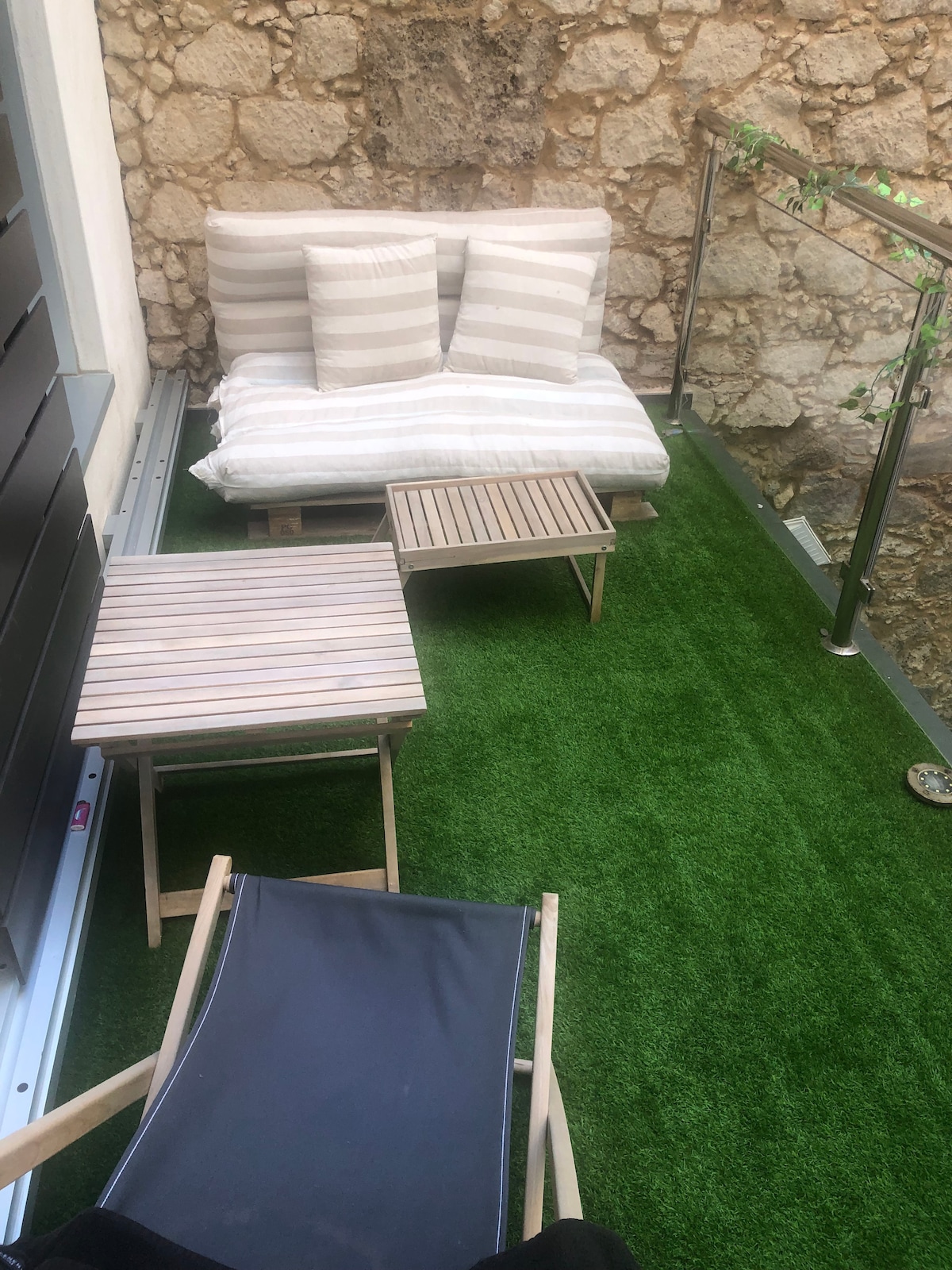 A small terrace is furnished with two lightweight wooden tables and a pair of seating options. One seating area features a cushioned sofa adorned with striped pillows, while a single reclining chair offers a place to relax. The space is surrounded by stone walls and artificial grass.