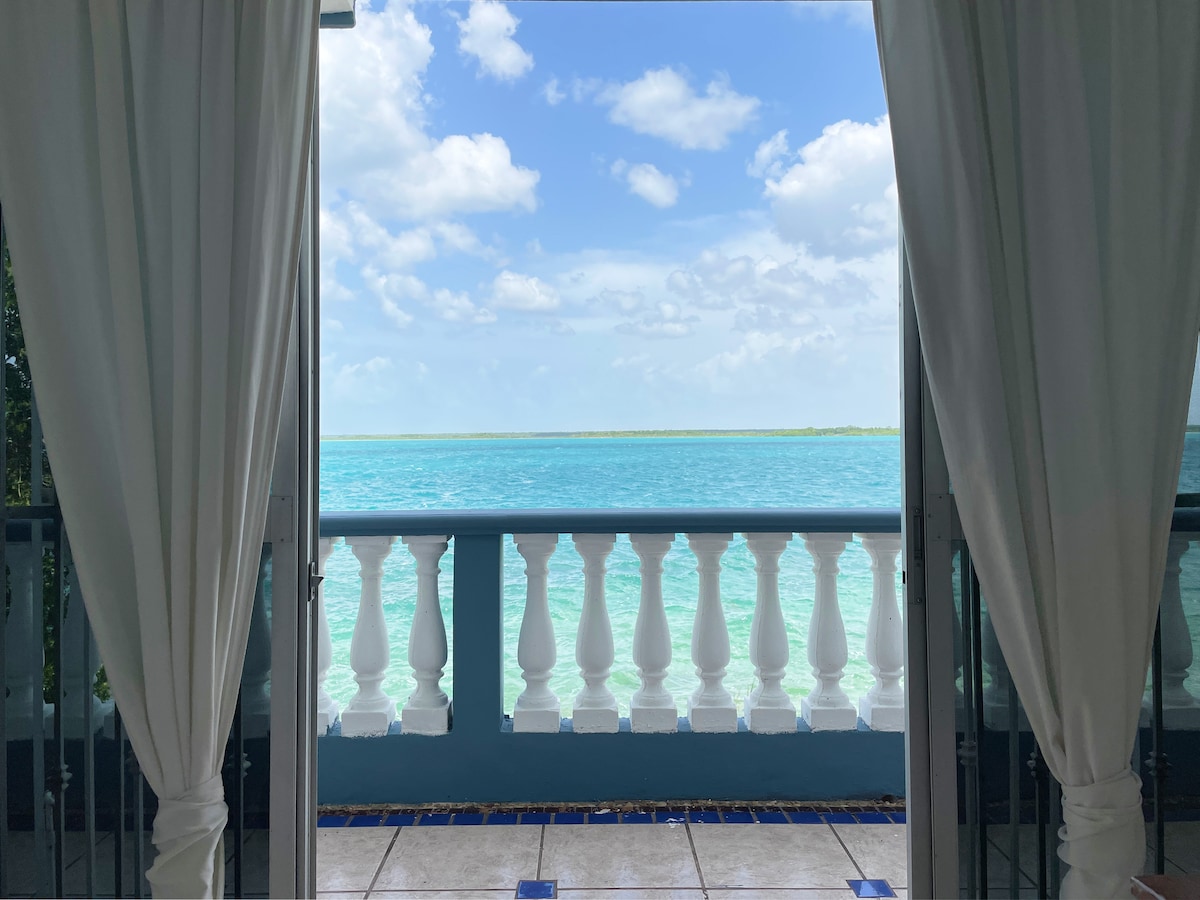 A serene view of the lagoon is framed by elegant white curtains. The vibrant turquoise water stretches toward the horizon under a sky scattered with fluffy clouds, creating a peaceful atmosphere for relaxation.