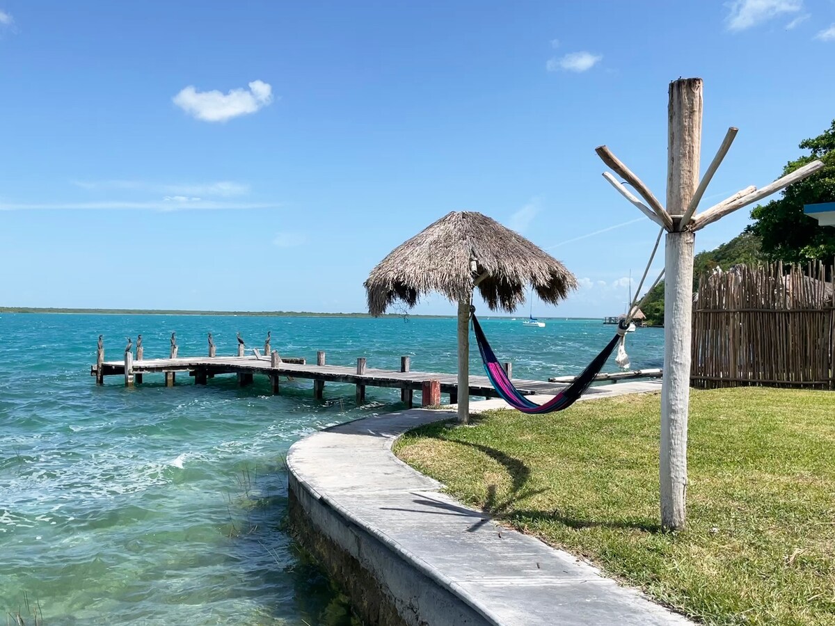 A view of the lagoon features a wooden dock extending into the water, shaded by a thatched-roof hut. A colorful hammock is suspended nearby, offering a relaxing spot. The water displays shades of blue and green, reflecting the clear sky.